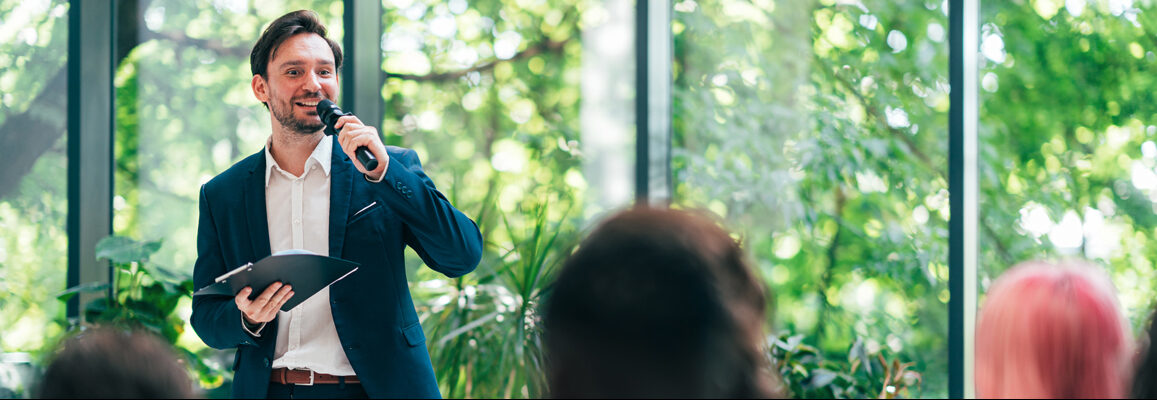 A speaker talking to a seated crowd while holding a microphone and some papers.